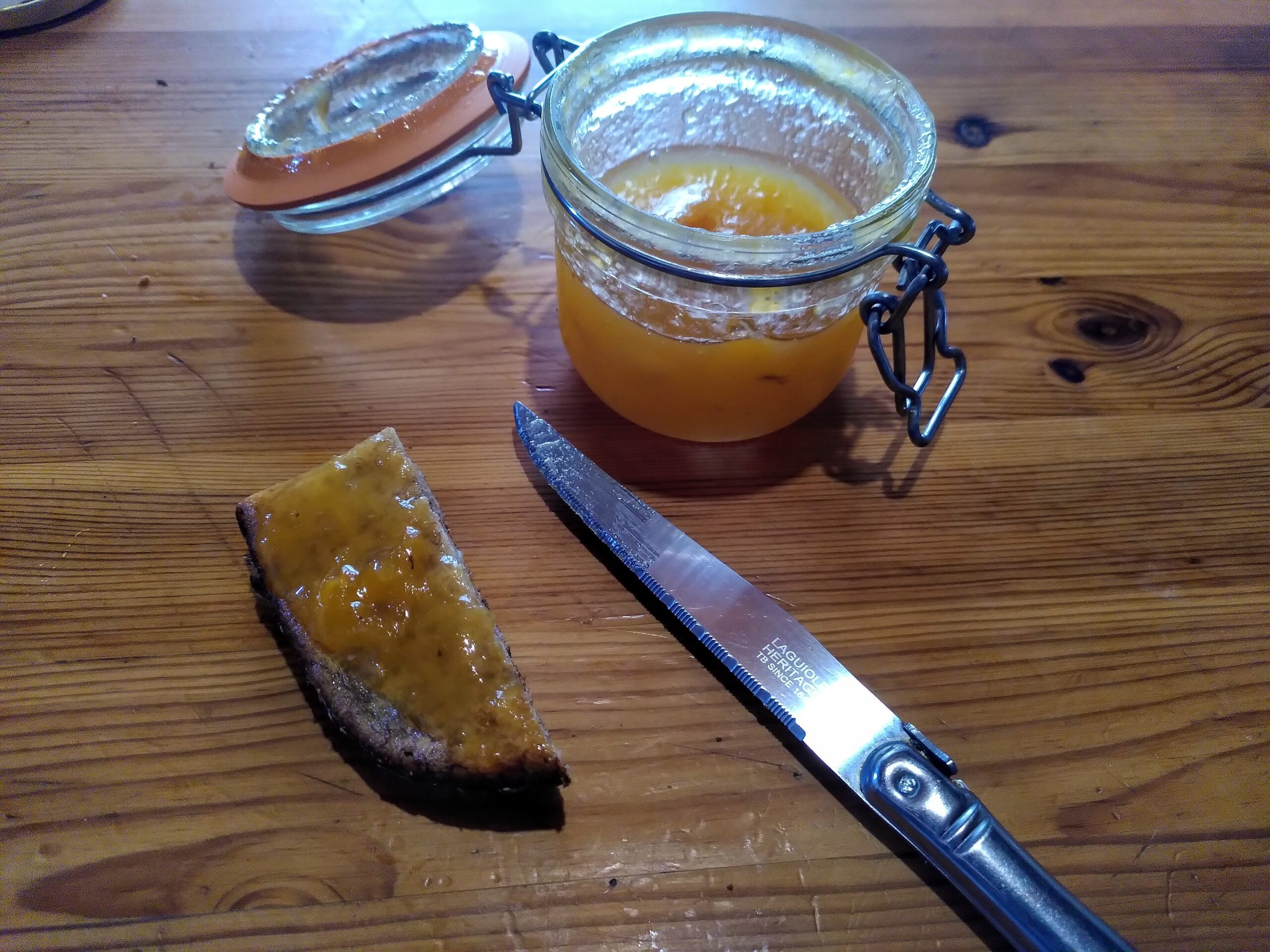 Un pot de confiture de kaki ouvert, posé sur une table. A côté se trouve un couteau et un gâteau qui est tartiné de cette confiture.