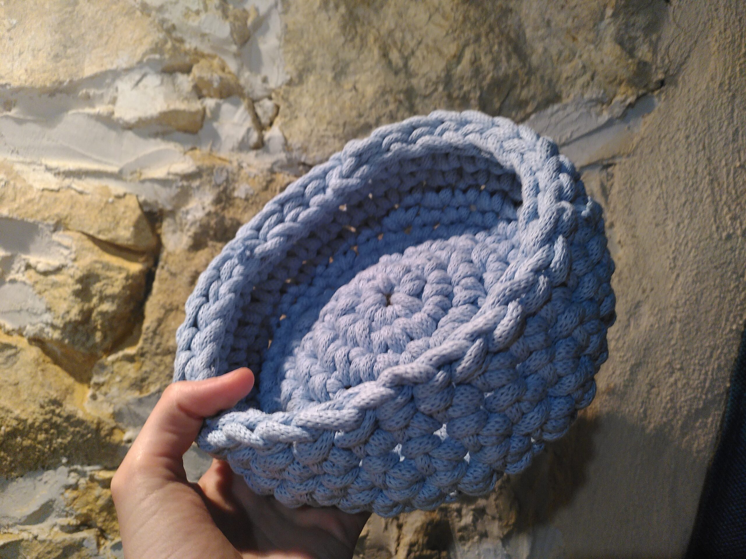 Une panière bleue, faite en crochet. Elle est tenue par une main, face à un mur.