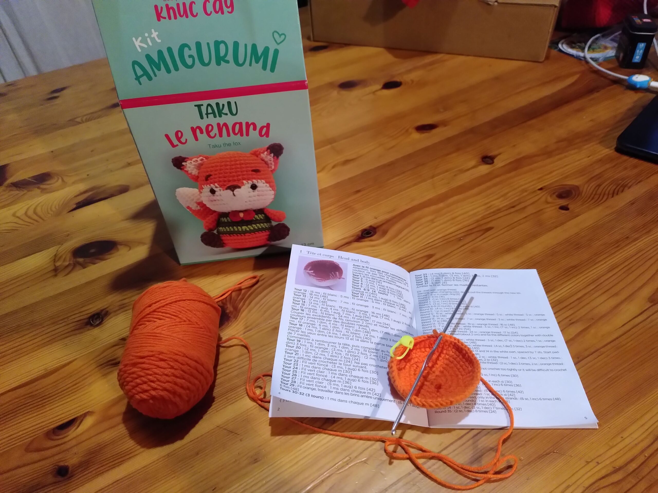 Une boite avec inscrit "Kit amigurumi :Taku le renard". À côté ce trouve le manuel pour faire ce renard, une pelote de laine orange, et le début du renard, et un crochet.